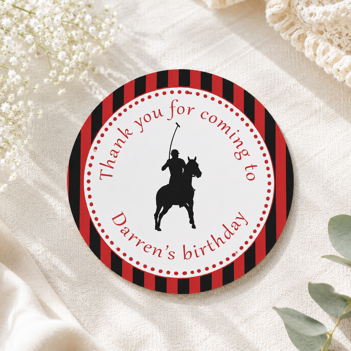 A round printed favor tag or sticker with red and black striped border, a black polo player silhouette, and the text 'Thank you for coming to Darren's birthday,' styled on cream fabric with baby's breath and eucalyptus.